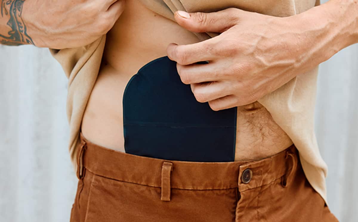 Mann mit schwarzem Stomabeutel von Coloplast am Bauch hebt sein beiges T-Shirt nach oben