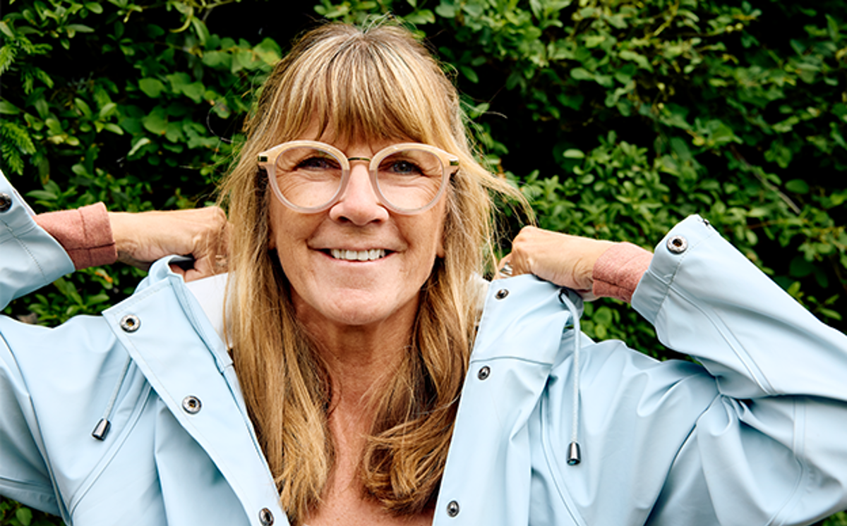 Frau blonden Haaren und blauer Regenjacke lächelt und setzt Kapuze auf